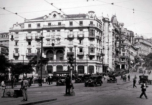 Fotografia antiga de edifício de esquina, com bondes e automóveis de época