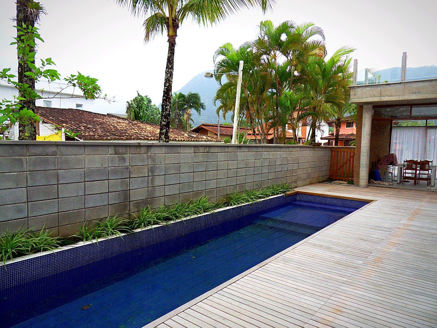 Piscina revestida de pastilhas azuis, deck de madeira e muro de blocos com forração