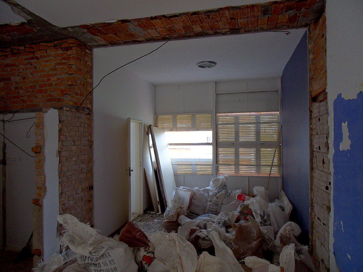 Paredes demolidas e sacos de entulho na sala, com venezianas ao fundo