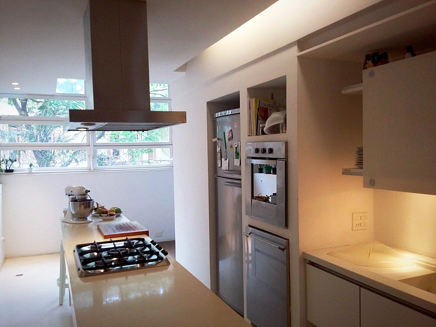 Cozinha com bancada branca, cooktop, coifa central e caixilho para a rua