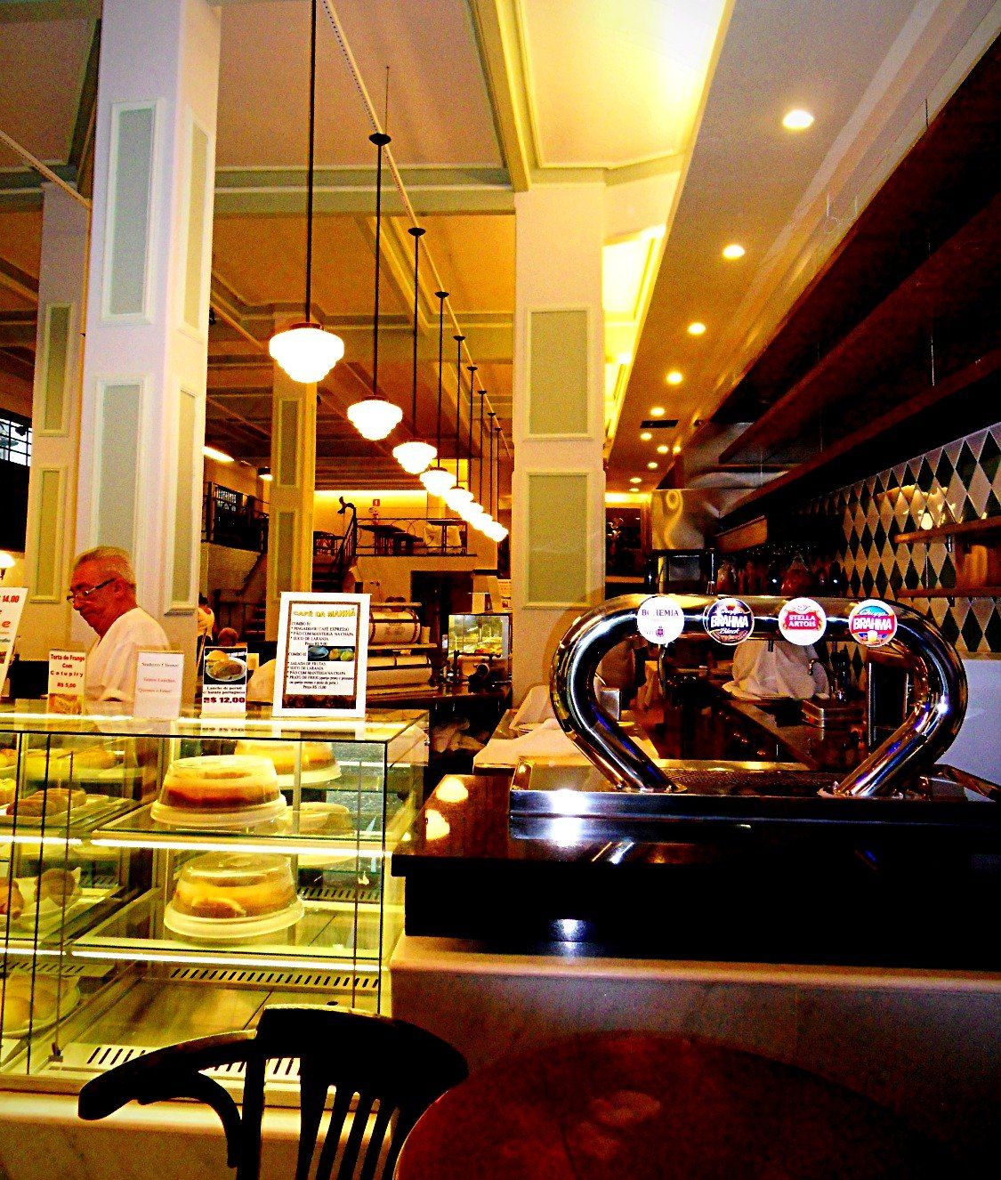 Chopeira cromada sobre o balcão, vitrine de doces e luminárias pendentes ao fundo